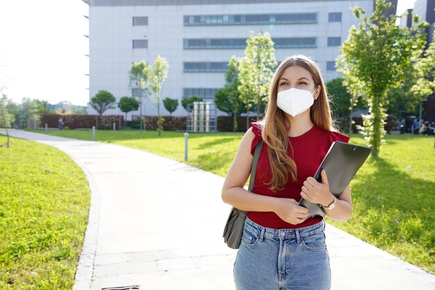 La ragazza di affari con la maschera protettiva cammina tenendo le cartelle nelle sue mani con edifici per uffici sullo sfondo