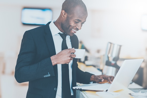 Business on the go. Cheerful young African man in formalwear using his laptop while standing at the bar