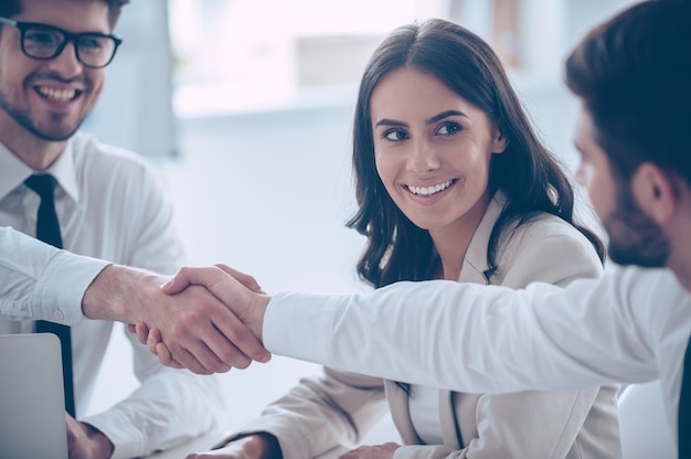 Foto stretta di mano di affari. primo piano di due giovani che si stringono la mano con un sorriso mentre sono seduti in ufficio con il loro bellissimo collega