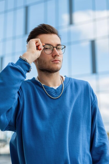 Business handsome man in blue trendy sweatshirt wear a fashion glasses and walks in the city near a business building