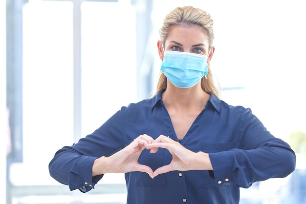 Foto cuore d'affari e ritratto di una donna con cure covid ed emoji per una pandemia sul lavoro sicurezza sanitaria e dipendente aziendale con un gesto della mano d'amore mentre indossa una maschera durante la corona