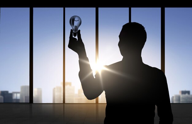 business, inspiration, idea and people concept - silhouette of businessman holding light bulb over office window background