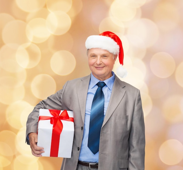 business, kerst, cadeaus en mensen concept - lachende senior man in pak en kerstmuts met cadeau over beige lichten achtergrond