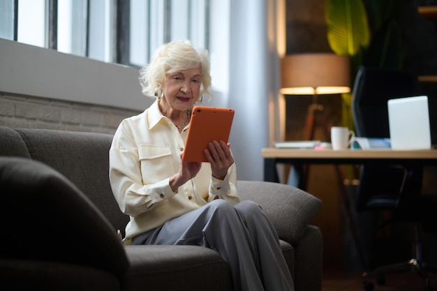 Business lady. gray-haired mature elegant woman working on
tablet sitting on sofa near window in modern room during
daytime