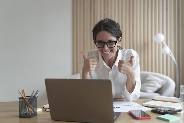 Деловая леди смотрит на экран компьютера с широкой улыбкой, показывая большой палец обеими руками