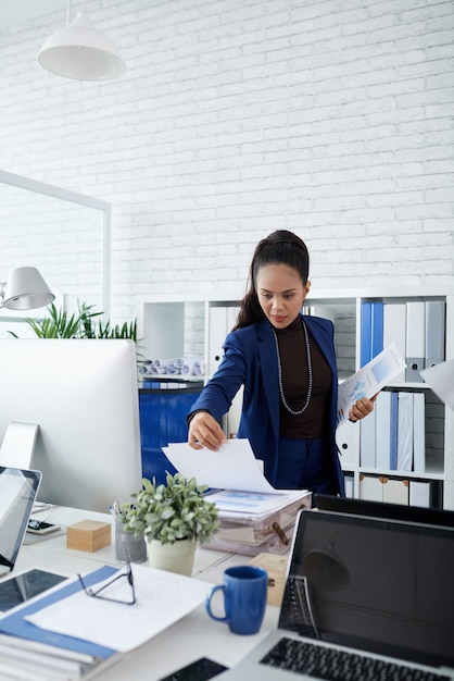 Business lady taking documents with charts and reports from stack next to her computer