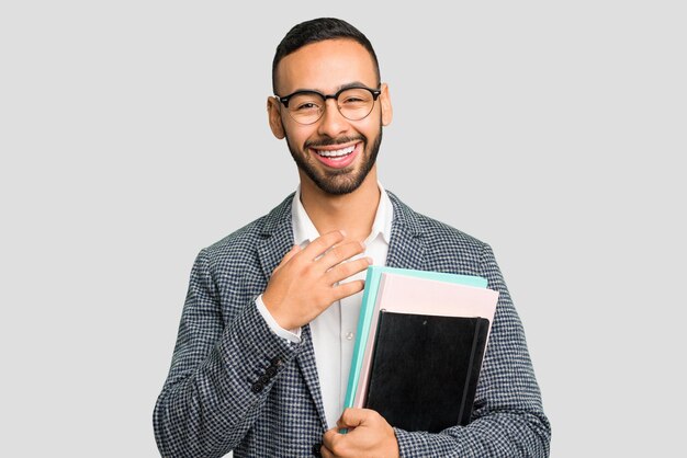 Foto uomo d'affari latino che tiene libri isolato ride ad alta voce tenendo la mano sul petto