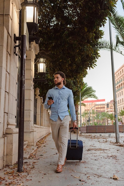スーツケースと路上で携帯電話を持つビジネス ラテン男