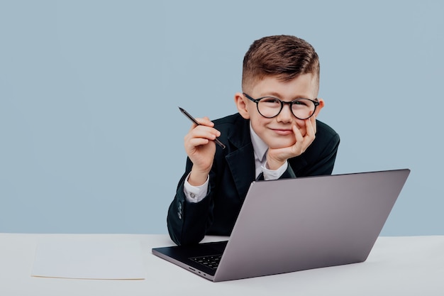 Business little boy in glasses and suit reading data from laptop looks at camera sitting at table an...