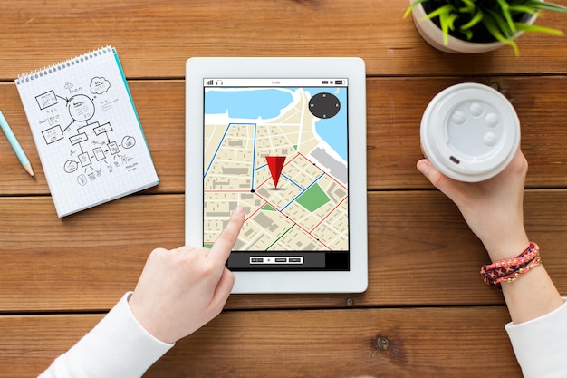 business, location, education, technology and people concept - close up of woman with gps navigator map on tablet pc computer screen and coffee on wooden table with gps navigator map on screen