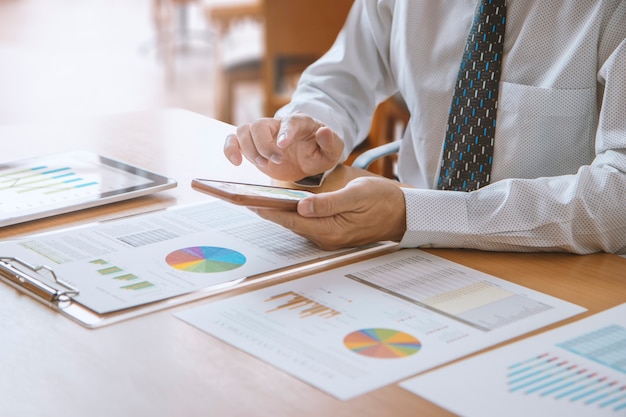 Business man or analyst in a modern office touching smartphone\
while reviewing financial statements on business performance or\
return on investment