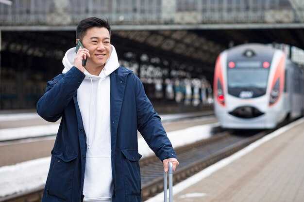 Uomo d'affari uomo asiatico alla stazione ferroviaria divertendosi a parlare al telefono un passeggero è arrivato in visita d'affari in una nuova città con una grande valigia di bagagli