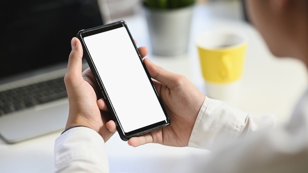 business man holding on crop black smartphone with white isolated blank screen with the blurred working desk