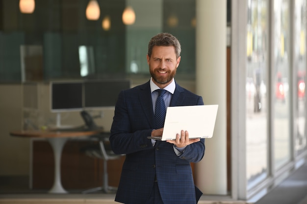 Деловой человек, держащий свой ноутбук, стоя в костюме на фоне офиса