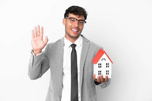 Business man holding a house isolated on blue background saluting with hand with happy expression