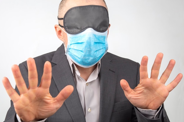 Business man in a medical mask and blindfold for sleeping with hands up on a white