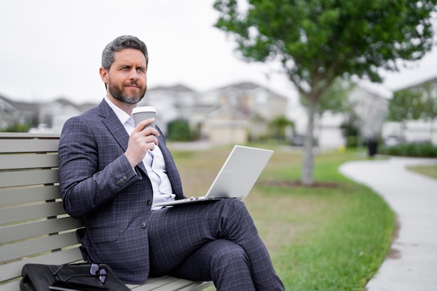 Business man sitting on bench in park work on laptop drink coffee businessman online work from lapto