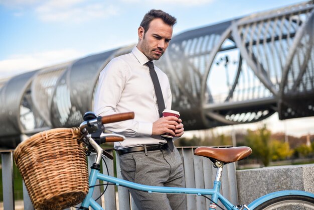 コーヒーを飲んで彼のビンテージ自転車のそばに立っているビジネスマンxA