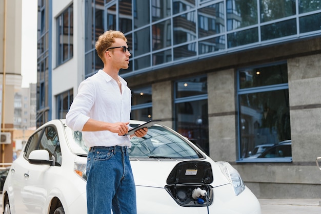 電気自動車の充電の近くに立って、通りでタブレットを使用しているビジネスマン。
