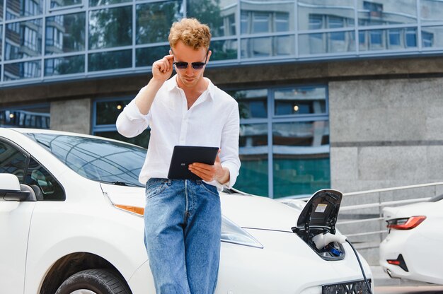 電気自動車の充電の近くに立って、通りでタブレットを使用しているビジネスマン。