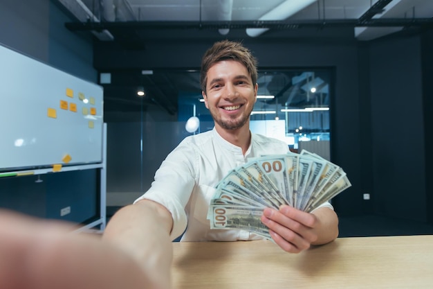 Business man talking on video communication holding phone joyfully showing cash dollars smiling and joking taking selfies on camera