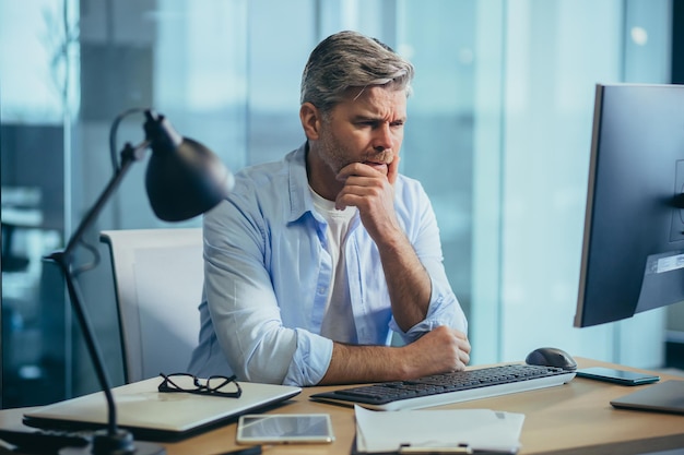 Photo business man in thought solves important issues and makes choices grayhaired businessman works at computer in modern office