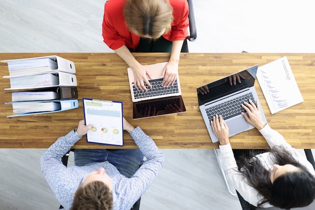 Business man and two women are sitting at wooden table with laptops and documents in office. Organization of negotiations concept.