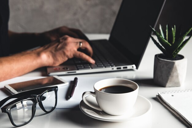 Business man using laptop computer and hand typing on laptop keyboard with notebook pen glasses and cup of hot coffee