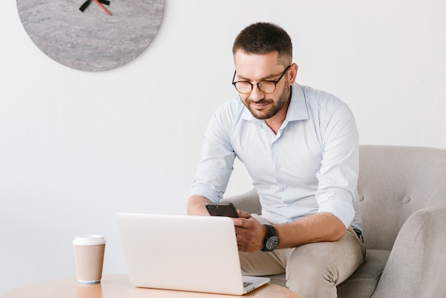 business man wearing eyeglasses sitting in armchair, and using smartphone while working on laptop in office interior