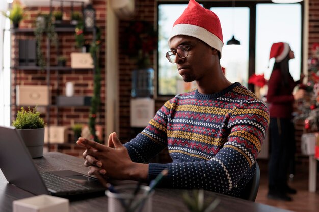 Business man working on laptop in startup office, using computer in festive workplace decorated with christmas tree and holiday ornaments. Male worker at company job during winter season.