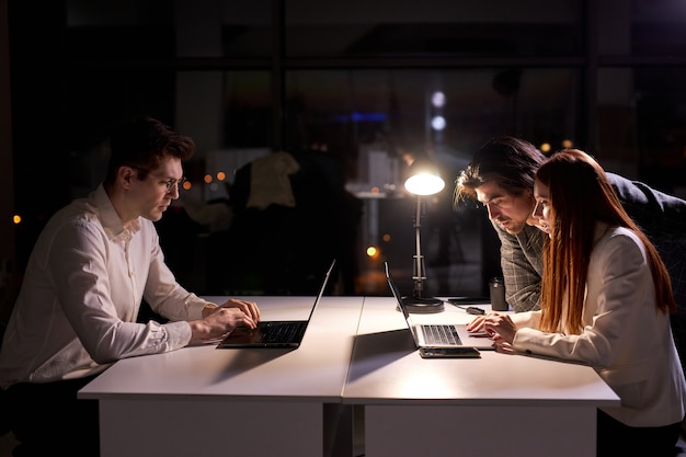 Business manager and employees work together on project, using laptop in office at late night, brainstorming, thinking a lot, solving tasks and questions. side view on office workers in formal wear