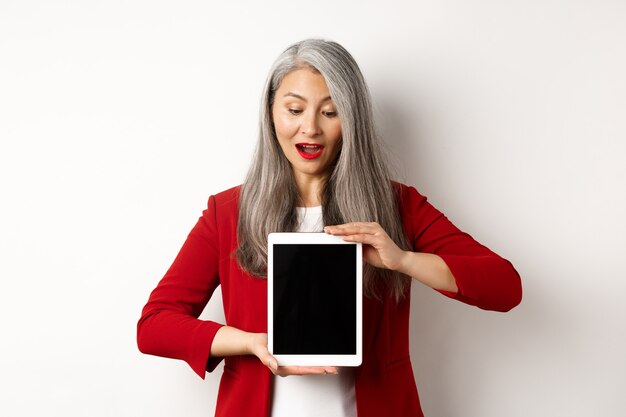 Business. Mature asian office lady showing digital tablet screen and looking down amazed, standing over white background.