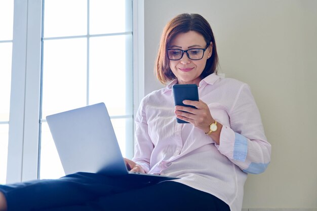 Business mature woman sitting on floor at home using laptop and smartphone