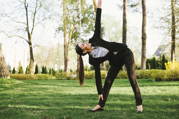 Business and meditation A concentrated beautiful business woman in a black suit is sitting in a lotus position on the grass in the city park and resting after work