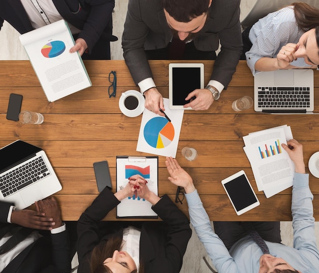 Incontro di lavoro. le persone impegnate lavorano in ufficio, vista dall'alto sul tavolo di legno con telefoni cellulari, laptop, tablet e documenti con diagramma, background aziendale