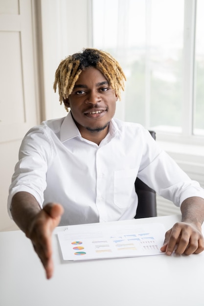 Business meeting Happy black man Greeting gesture Handsome stylish guy sitting office desk with documents proposing to shake hands in light room interior