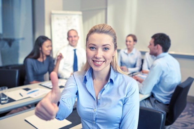 business, mensen, gebaar en teamwork concept - glimlachende zakenvrouw duimen opdagen met een groep zakenmensen die op kantoor bijeenkomen