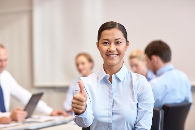 business, mensen, gebaar en teamwork concept - glimlachende zakenvrouw duimen opdagen met een groep zakenmensen die op kantoor bijeenkomen