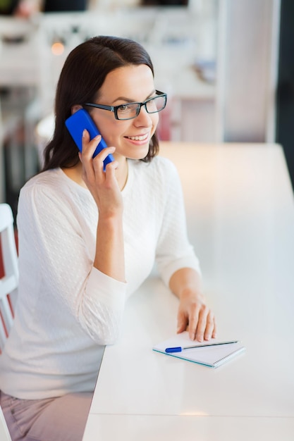 business, mensen, technologie en lifestyle concept - lachende jonge vrouw in brillen bellen op smartphone in cafe