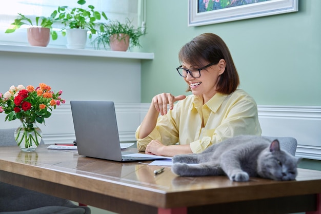 Business middle aged woman working from home using laptop along with pet cat Female sitting at table with documents sleeping cat lying next to owner Freelance remote work 40s people concept