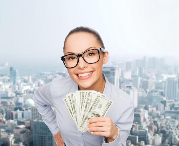 business, money, city and banking concept - smiling businesswoman in eyeglasses with dollar cash money over cityscape background