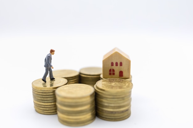 Business, Money, Finance, Home loan and management concept. Close up of businessman miniature figure walking on top of stack of gold coins to mini wooden house toy