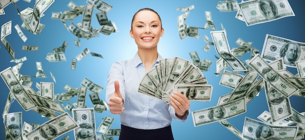 business, money, finance, people and banking concept - smiling businesswoman with heap of dollar cash money showing thumbs up over blue background