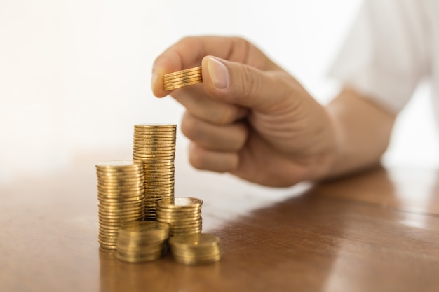 Business, Money, Finance, Secure and Saving Concept. Close up of man hand holding and put five coins to top of stack of gold coins on wooden table.