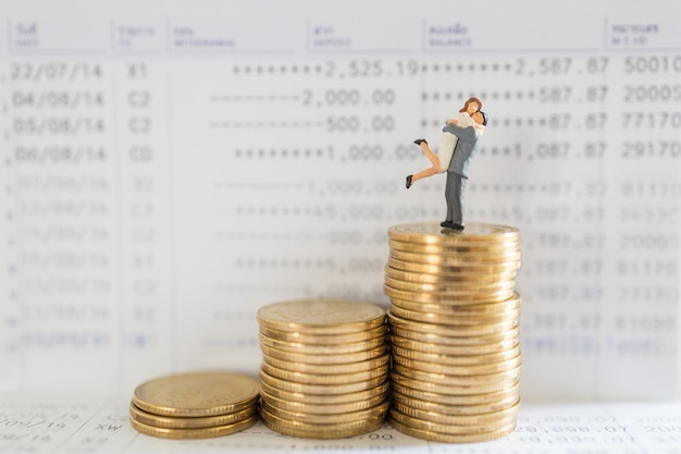 Business, Money, Saving, Security and Couple Family Concept. Close up of man and woman miniature figure people hug and standing on top of stack of gold coins on bank passbook with copy space.