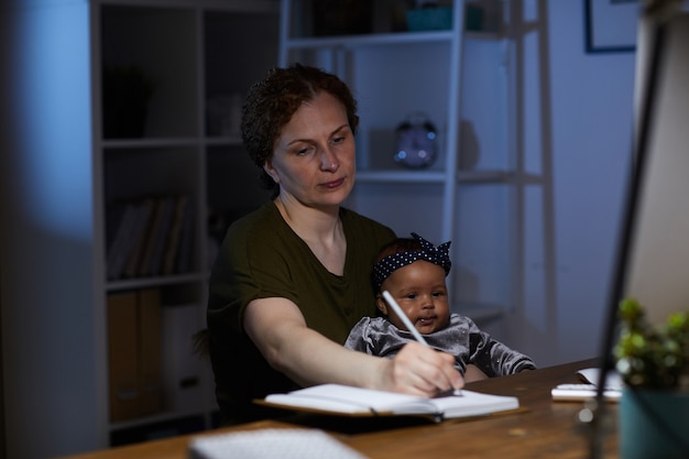 그녀는 밤까지 일하는 아기와 함께 그녀의 직장에 앉아있는 동안 노트 패드에 메모를 만드는 비즈니스 어머니