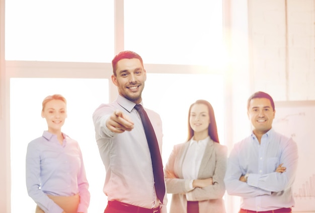 business and office concept - smiling handsome businessman with team in office pointing finger at you