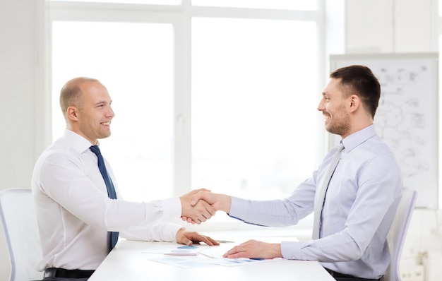концепция бизнеса и офиса - два улыбающихся бизнесмена пожимают друг другу руки в офисе