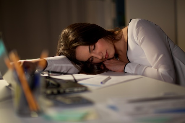 사진 사업, 과로, 마감일, 사람 개념 - 노트북과 서류를 가진 피곤한 여성이 밤 사무실에서 테이블 위에서 잠을 자고 있습니다.