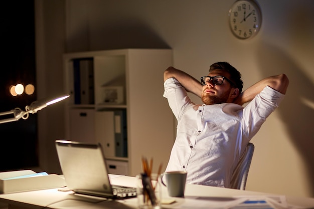 Photo business, overwork, deadline and people concept - tired man with laptop working at night office and stretching